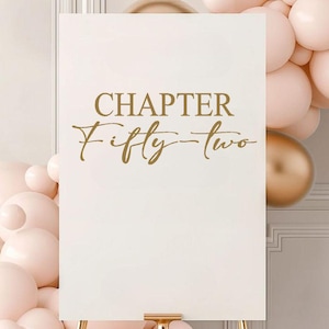 May include: A white sign with gold lettering that reads "CHAPTER Fifty-two." The sign is displayed on a gold stand, with pink and gold balloons in the background. The sign is likely for a birthday celebration.