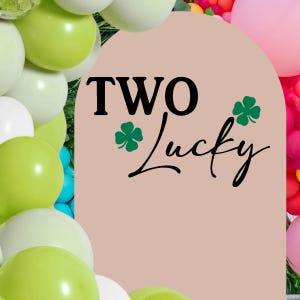 May include: A light pink arch-shaped sign with the words "TWO Lucky" in black script and green shamrock accents. The sign is surrounded by a colorful arrangement of balloons in shades of green, white, pink, and blue.