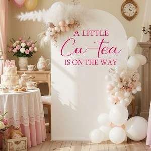 May include: A large white arched backdrop featuring the pink text A LITTLE Cu-Tea IS ON THE WAY, decorated with white and pale pink balloons and dried pampas grass. The display is set in a room with a table, a tiered cake, and floral arrangements.