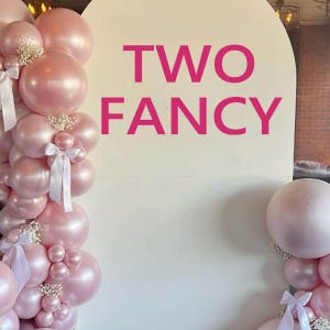 May include: A decorative display featuring pink balloons, white bows, and floral accents. The words "TWO FANCY" are displayed in bright pink text against a white backdrop, suitable for a party or event.