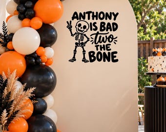 May include: A beige arched backdrop featuring a black graphic of a skeleton making a peace sign with the text "ANTHONY IS BAD TO THE BONE" and a balloon garland in orange, black, and white colors surrounding the left and top edges of the arch.
