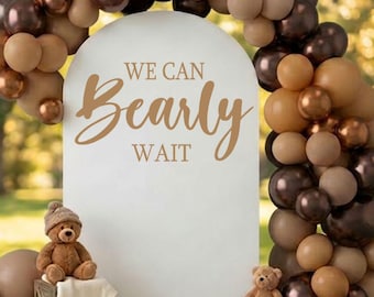 May include: A white arched backdrop featuring the text WE CAN Bearly WAIT in brown lettering, surrounded by a balloon garland in shades of tan, beige, and metallic brown. Two brown plush teddy bears are positioned on wooden crates at the base.