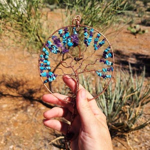 May include: A copper wire tree of life pendant, adorned with turquoise and amethyst gemstones. The tree design is circular, with branches and roots crafted from the copper wire. The pendant is held in front of a blurred natural background.