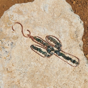 May include: Copper wire cactus pendant with green and blue stone accents. The cactus design features a hook at the top for hanging. The pendant is displayed on a light-colored stone surface.