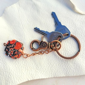 May include: A copper key chain with a red and black patterned charm, a copper clasp, and a copper ring attached to two keys.