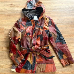 May include: A hooded jacket constructed from a patchwork of multi-colored leather pieces in shades of brown, red, black, and tan. The garment features a front button closure, long sleeves, and a matching leather tie belt at the waist.