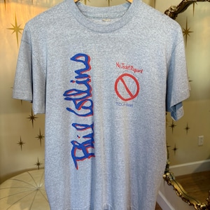 Op de afbeelding: Een grijs t-shirt met de naam "Phil Collins" in blauwe en rode verticale tekst. Het shirt heeft ook de tekst "No Jacket Required" boven een rode cirkel met een ontwerp van een doorgestreepte fietswiel. Het t-shirt hangt aan een houten hanger.