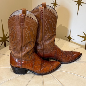 May include: A pair of brown leather cowboy boots with intricate stitching and a textured, crocodile-like pattern on the foot and upper shaft. The boots have pointed toes and stacked heels, showcasing a classic Western style.