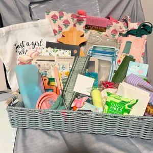 May include: A rectangular gray woven basket filled with various items including a white tote bag with the text Just a girl who, a pink floral shopper tote, a green cucumber face wipe package, a blue silicone pouch, and assorted stationery.