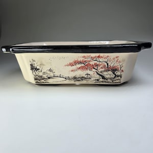 May include: Rectangular ceramic bonsai pot with a black rim and feet. The off-white sides feature a hand-painted Asian landscape with a bridge, trees, and red foliage. The pot is designed for growing small plants.