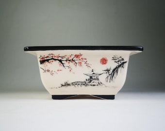 May include: Rectangular bonsai pot with a black rim and base. The off-white ceramic pot features a hand-painted Japanese landscape with a red sun, cherry blossoms, and a pagoda. The pot is designed for growing small trees.