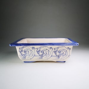 Può includere: Un vaso rettangolare in ceramica bianca con un motivo a onde blu e bianco. Il vaso ha un bordo leggermente rialzato e tre piccoli piedini.