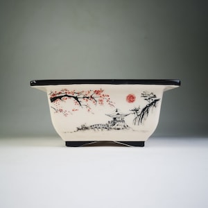 Puede incluir: Maceta rectangular para bonsái con borde y base negros. La maceta de cerámica blanquecina presenta un paisaje japonés pintado a mano con un sol rojo, flores de cerezo y una pagoda. La maceta está diseñada para cultivar árboles pequeños.