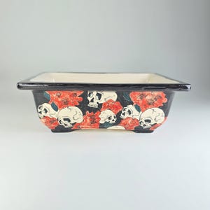 Pode incluir: Um vaso de cerâmica preto com uma borda branca e um design de caveiras e flores. O vaso tem uma forma quadrada e um fundo plano.
