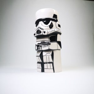 May include: A white ceramic tiki mug shaped like a Star Wars Stormtrooper. The mug has black details and a black blaster rifle.