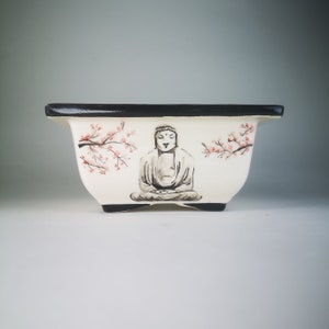 Może przedstawiać: Biała ceramiczna doniczka z czarną krawędzią. Doniczka ma ręcznie malowany wzór medytującej postaci Buddy z gałązką wiśni po obu stronach.