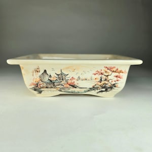 Peut inclure: Un pot à bonsaï en céramique blanche avec une scène peinte d'une pagode, d'arbres et d'un lac. Le pot a une forme carrée avec des bords arrondis et quatre petits pieds.