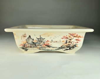 May include: A white ceramic bonsai pot with a painted scene of a pagoda, trees, and a lake. The pot has a square shape with rounded edges and four small feet.
