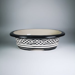 Puede incluir: Una maceta ovalada de cerámica negra y blanca con un diseño de nudo celta. La maceta tiene un acabado liso y esmaltado.