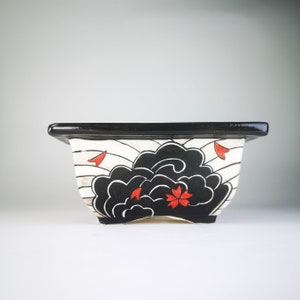 May include: Black and white ceramic planter with a stylized wave design and red flower accents. The planter has a black rim and a white interior.