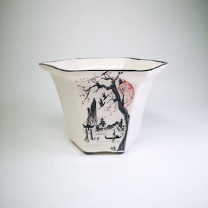 Puede incluir: Maceta de cerámica blanca con un diseño inspirado en japonés en negro y rojo. El diseño presenta un árbol con flores rojas, una montaña, una pagoda y un barco.