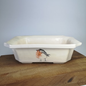May include: Cream-coloured rectangular ceramic baking dish with a hand-painted robin bird design. The bird has a red breast, grey wings, and a black head. The dish has a slightly raised rim and is set on a wooden surface.