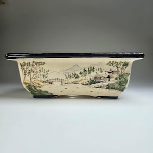 May include: A rectangular ceramic bonsai pot with a cream-colored base and a black rim. The exterior features a hand-painted landscape illustration depicting a bridge, traditional buildings, trees, and mountains in shades of green and brown.