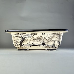 Peut inclure: Un pot rectangulaire en céramique avec un bord noir et un fond blanc. Le pot présente un dessin à l'encre noir et blanc d'un paysage japonais avec des montagnes, des arbres, un pont et une pagode.
