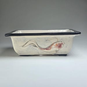 Pode incluir: Jardineira retangular de cerâmica com base creme e borda preta. A jardineira apresenta um desenho de peixe koi desenhado à mão em tons de vermelho, laranja e preto. A jardineira tem cerca de 25 cm de comprimento.