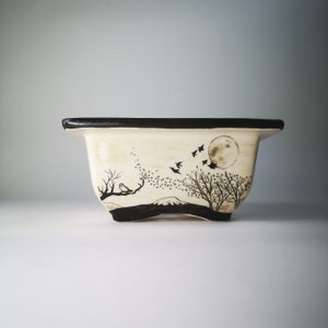 May include: A white ceramic planter with a black rim and a black and white design of birds flying over a moon and trees.