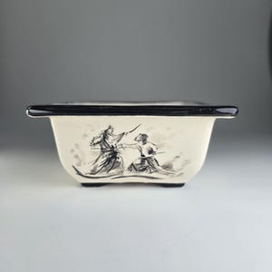 Könnte beinhalten: Ein quadratischer Bonsai-Topf aus weißer Keramik mit schwarzem Rand und einer Schwarzweiß-Tuschzeichnung von zwei Figuren in Kampfstellung.