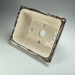 May include: A white ceramic planter with a black rim and drainage holes. The planter is rectangular and has a smooth, glossy finish.