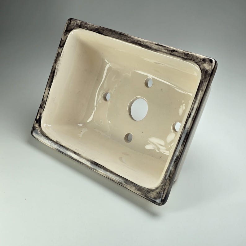 May include: A white ceramic planter with a black rim and drainage holes. The planter is rectangular and has a smooth, glossy finish.