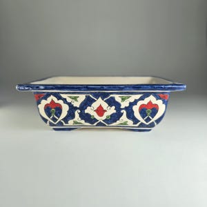 Könnte beinhalten: Ein quadratischer Keramik-Pflanztopf mit einem blauen und weißen geometrischen Muster. Der Topf hat einen weißen Innenraum und einen blauen Außenraum mit roten und grünen Akzenten.