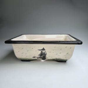 May include: Rectangular ceramic bonsai pot with a cream-colored interior and a black rim. The exterior features a black and white illustration of a samurai in the rain. The pot has a low profile and is designed for bonsai trees.