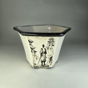 Op de afbeelding: Witte keramische plantenbak met een zwarte rand en een zwart-witte illustratie van een samoerai die voor een pagode staat.