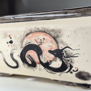 May include: A white ceramic bonsai pot with a black and white ink drawing of a dragon curled around a red circle. The pot has a black rim and a speckled surface.