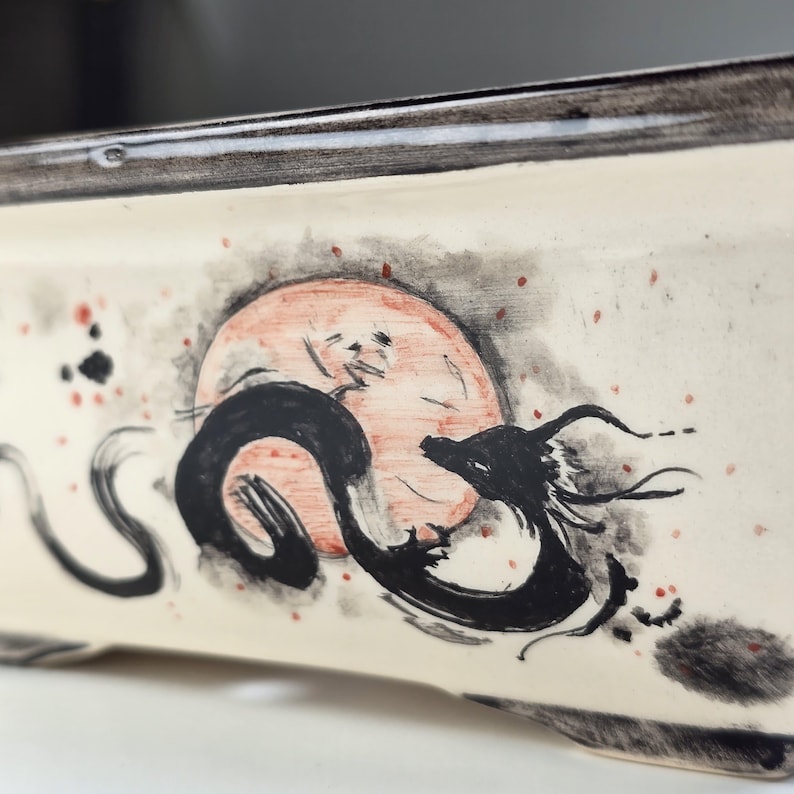 May include: A white ceramic bonsai pot with a black and white ink drawing of a dragon curled around a red circle. The pot has a black rim and a speckled surface.