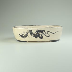 May include: A white oval ceramic bonsai pot with a black dragon design. The pot has a black rim and a black base.