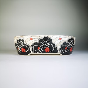 May include: A white ceramic bonsai pot with a black and red design of stylized clouds and flowers. The pot has a rounded oval shape and a slightly raised rim.