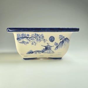 Könnte beinhalten: Rechteckiger Bonsai-Topf aus Keramik mit cremefarbenem Boden und dunkelblauem Rand. Der Topf hat ein handgemaltes blau-weißes Landschaftsdesign mit einer Pagode, einer Brücke und Bäumen. Der Topf hat drei kleine Füße.