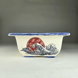 May include: A white ceramic rectangular planter with a blue rim and a painted design of a red sun and blue waves.