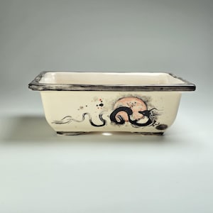 May include: A white ceramic rectangular bonsai pot with a black dragon design and a red and orange moon. The pot has a black rim and a drainage hole.