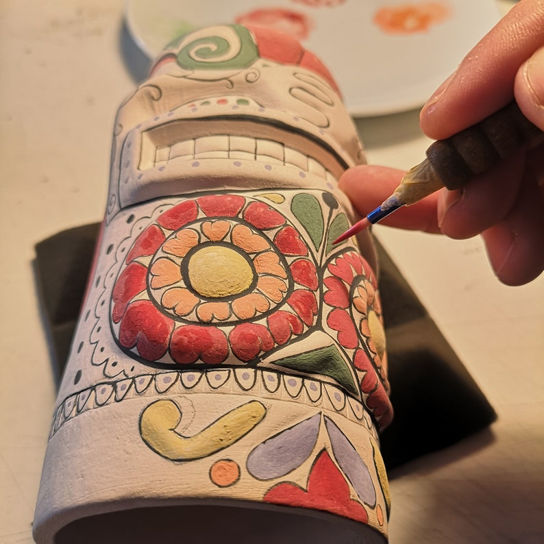 May include: A white ceramic skull with colorful floral designs is being painted with a fine-tipped brush. The skull has a toothy grin and is decorated with red, green, yellow, and purple paint.