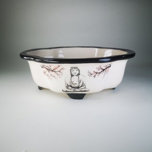 May include: White ceramic planter with a black rim and three legs. The planter features a hand-painted design of a meditating figure with pink cherry blossoms.