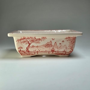 Könnte beinhalten: Rechteckiger Keramik-Pflanztopf mit rot-weißem, asiatisch inspiriertem Landschaftsdesign. Der Pflanztopf zeigt eine detaillierte Illustration von Bäumen, Bergen, Brücken und Vögeln. Die Kanten sind leicht gebogen, und die Basis hat kleine Füße.