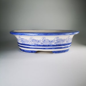 Op de afbeelding: Witte ovale keramische plantenbak met een blauw en wit golfontwerp. De plantenbak heeft een blauwe rand en blauwe strepen rond de onderkant.