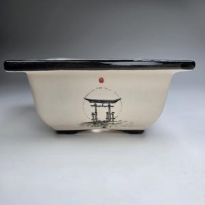 May include: Cream-coloured rectangular bonsai pot with a black rim and feet. The front features a black and white illustration of a Japanese torii gate with a red dot above it. The pot is designed for bonsai trees.