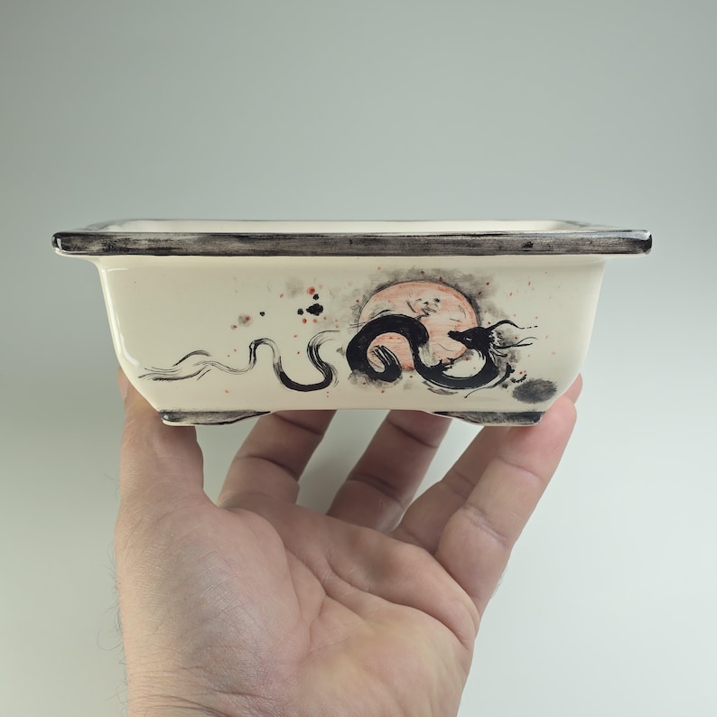 May include: A white ceramic bonsai pot with a black and white dragon design and a red and orange moon. The pot has a dark gray rim and three small feet.