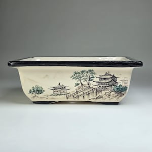 Może przedstawiać: Prostokątna ceramiczna doniczka do bonsai z kremową podstawą i grubą czarną krawędzią. Zewnętrzna strona ozdobiona jest ręcznie malowaną ilustracją krajobrazu w stylu tuszu, przedstawiającą tradycyjne pagody, most i drzewa w odcieniach czerni i zieleni.